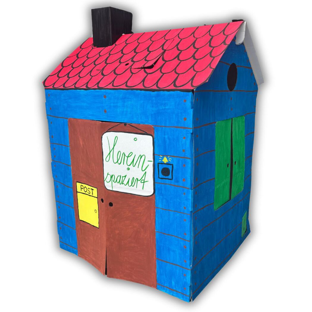 Großes, bunt bemaltes Spielhaus aus stabiler Wellpappe mit blauer Fassade, rotem Dach mit Ziegelmuster und brauner Tür. An den Seiten sind grüne Fenster aufgemalt, eine gelbe Postklappe sowie Klingel sind dargestellt. Ein Namensschild ist an der Tür befestigt.