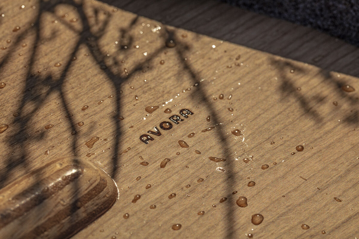 Nahaufnahme eines nassen Holzbretts mit der Aufschrift 'AVORA' in dunkler Gravur. Schatten von Ästen und Wassertropfen sind auf der Oberfläche sichtbar, was eine warme, natürliche und entspannte Atmosphäre schafft.