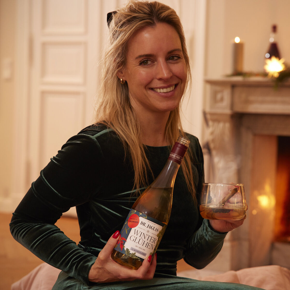 Eine Frau sitzt entspannt in einem gemütlichen Wohnzimmer vor einem brennenden Kamin. Sie trägt ein grünes Samtkleid, hält ein Glas und eine Flasche Winterglühen Riesling Winzerglühwein alkoholfrei von Dr. Jaglas. Im Hintergrund Dekoration, warme Beleuchtung und weihnachtliches Flair.