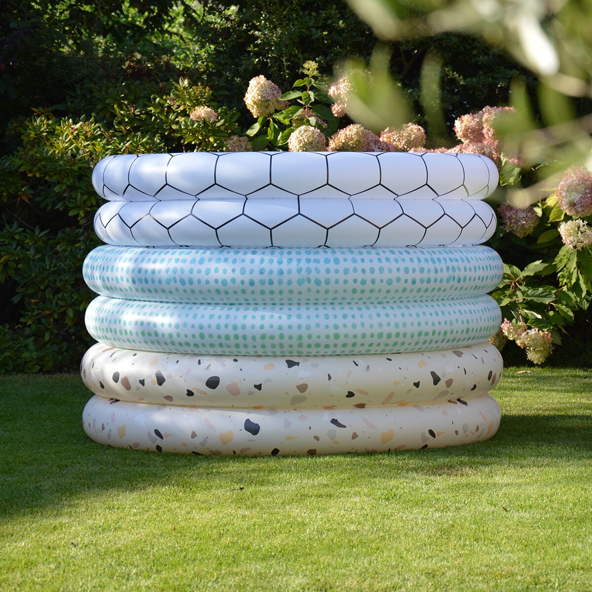 Im Vordergrund steht ein runder Swimmingpool aus weißem PVC mit unterschiedlichen Mustern: oben Waben, in der Mitte Punkte und unten Terrazzo-Optik. Der Pool steht auf grüner Rasenfläche, im Hintergrund blühende Hortensien und dichtes Grün.