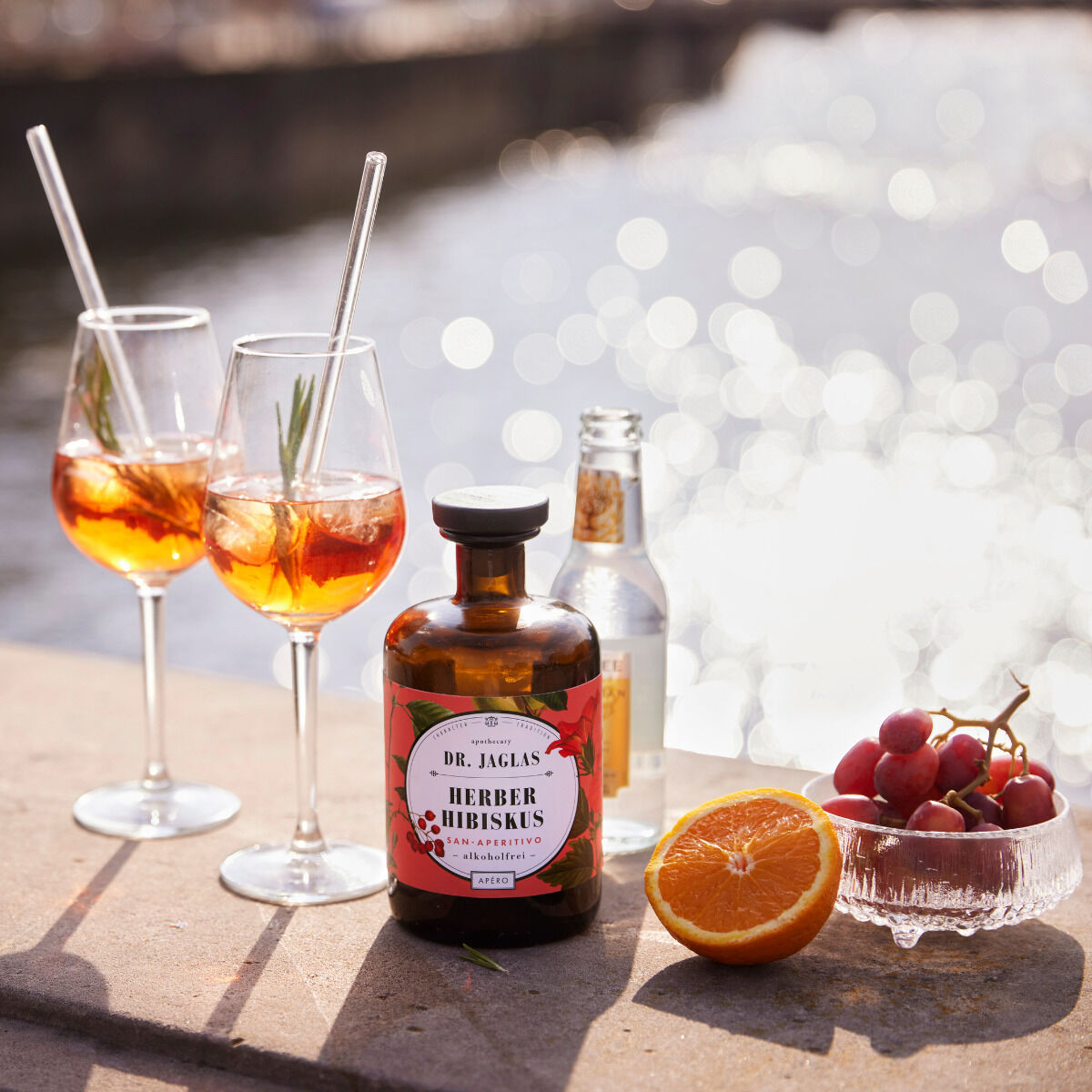 Flasche 'Herber Hibiskus' alkoholfreier Aperitif von Dr. Jaglas auf einem Tisch am Wasser, umgeben von zwei Cocktailgläsern, einer Flasche Mineralwasser, einer halben Orange und einem Glasschälchen mit roten Trauben. Sonnenlicht reflektiert auf dem Wasser im Hintergrund.