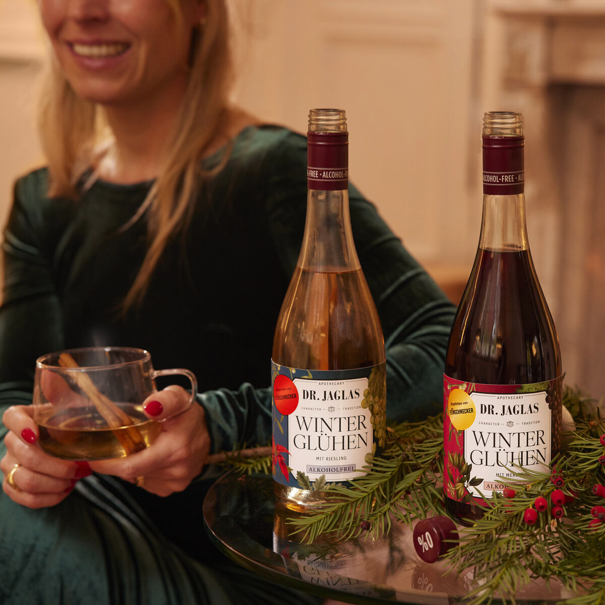 Eine Frau in einem dunkelgrünen Samtkleid sitzt entspannt vor einem Kaminfeuer. Auf einem Glastisch stehen zwei Flaschen Dr. Jaglas Winterglühen Rot + Weiß alkoholfrei, umgeben von Tannenzweigen mit roten Beeren. Zwei Gläser mit heißem Getränk betonen die winterlich-festliche Atmosphäre.