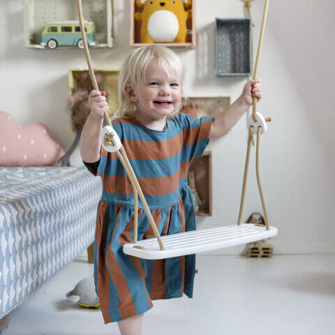 Ein kleines Kind sitzt in einem modern eingerichteten Kinderzimmer auf einer hölzernen Kinderschaukel mit Seilen, umgeben von spielerisch dekorierten Accessoires und Spielzeug. Die Schaukel ist für den Innenbereich konzipiert und lädt zum Spielen und Schwingen ein.