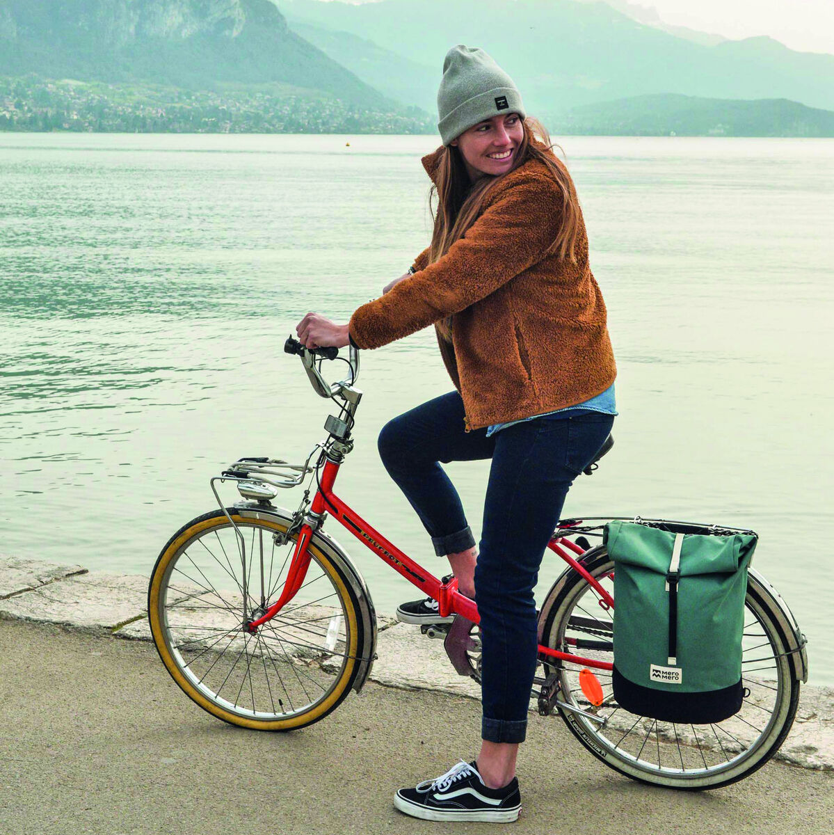 Eine Frau fährt ein Fahrrad mit einer angebrachten Fahrradtasche »Annecy« entlang eines Sees mit Berglandschaft im Hintergrund.