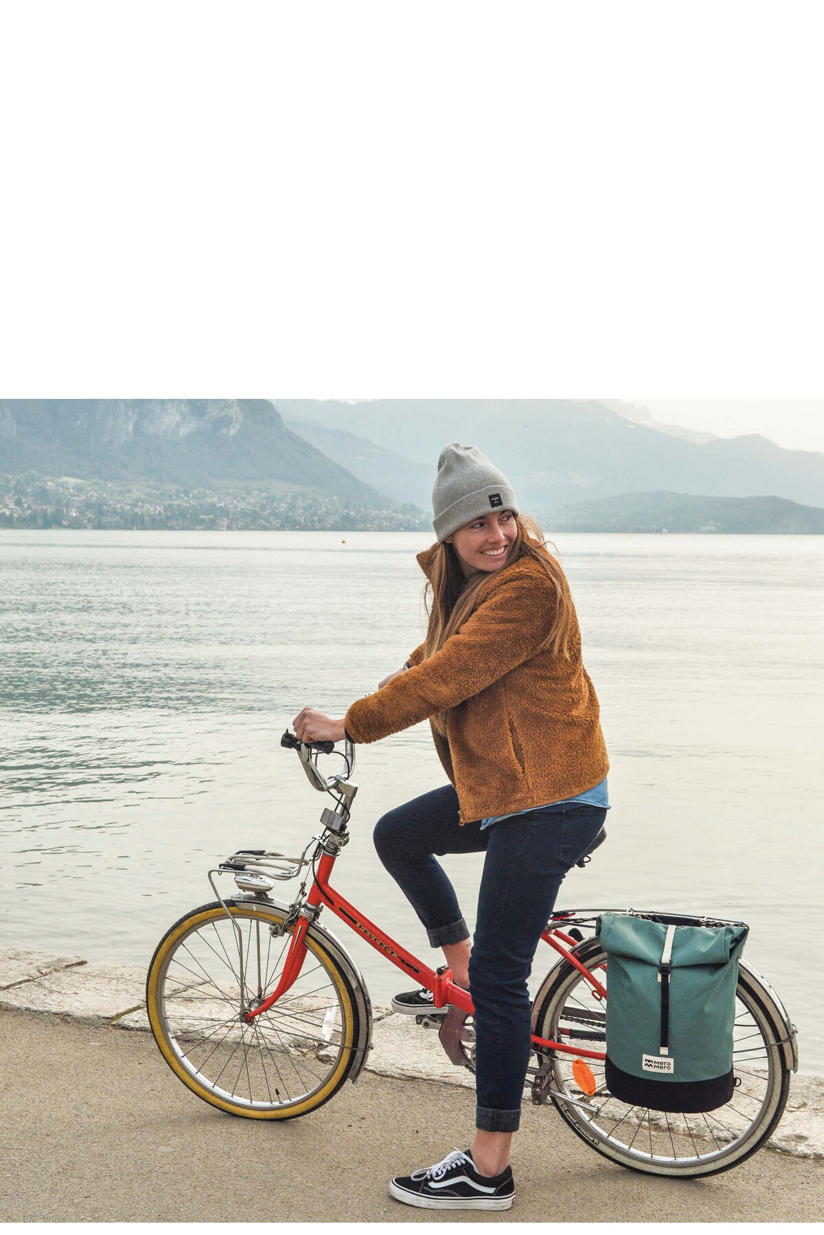 Eine Frau fährt ein Fahrrad mit einer angebrachten Fahrradtasche »Annecy« entlang eines Sees mit Berglandschaft im Hintergrund.