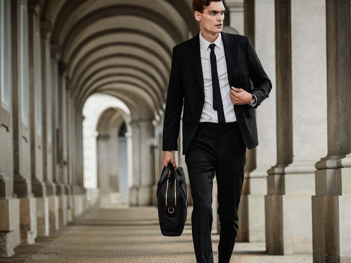 Ein Mann in einem dunklen Anzug und Krawatte hält die Businesstasche »ENDEAVOUR« von Mismo in der Hand und geht durch einen eleganten, überdachten Gang mit Bögen. Die Tasche ist schwarz und wirkt hochwertig.