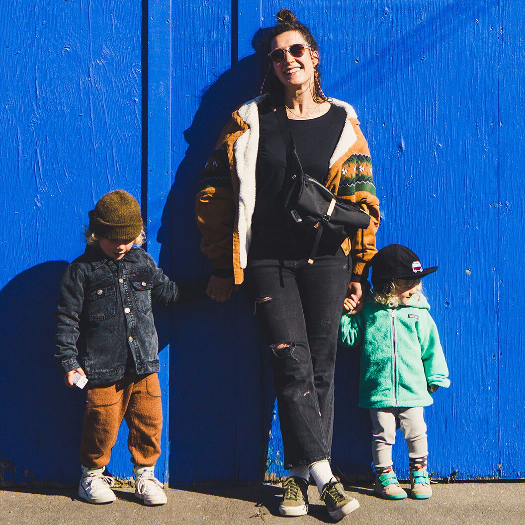 Eine Frau mit zwei Kindern steht vor einer blauen Wand. Sie trägt eine schwarze Tasche »Piha«. Die Frau ist modisch gekleidet und lächelt. Die Kinder tragen bunte Kleidung.