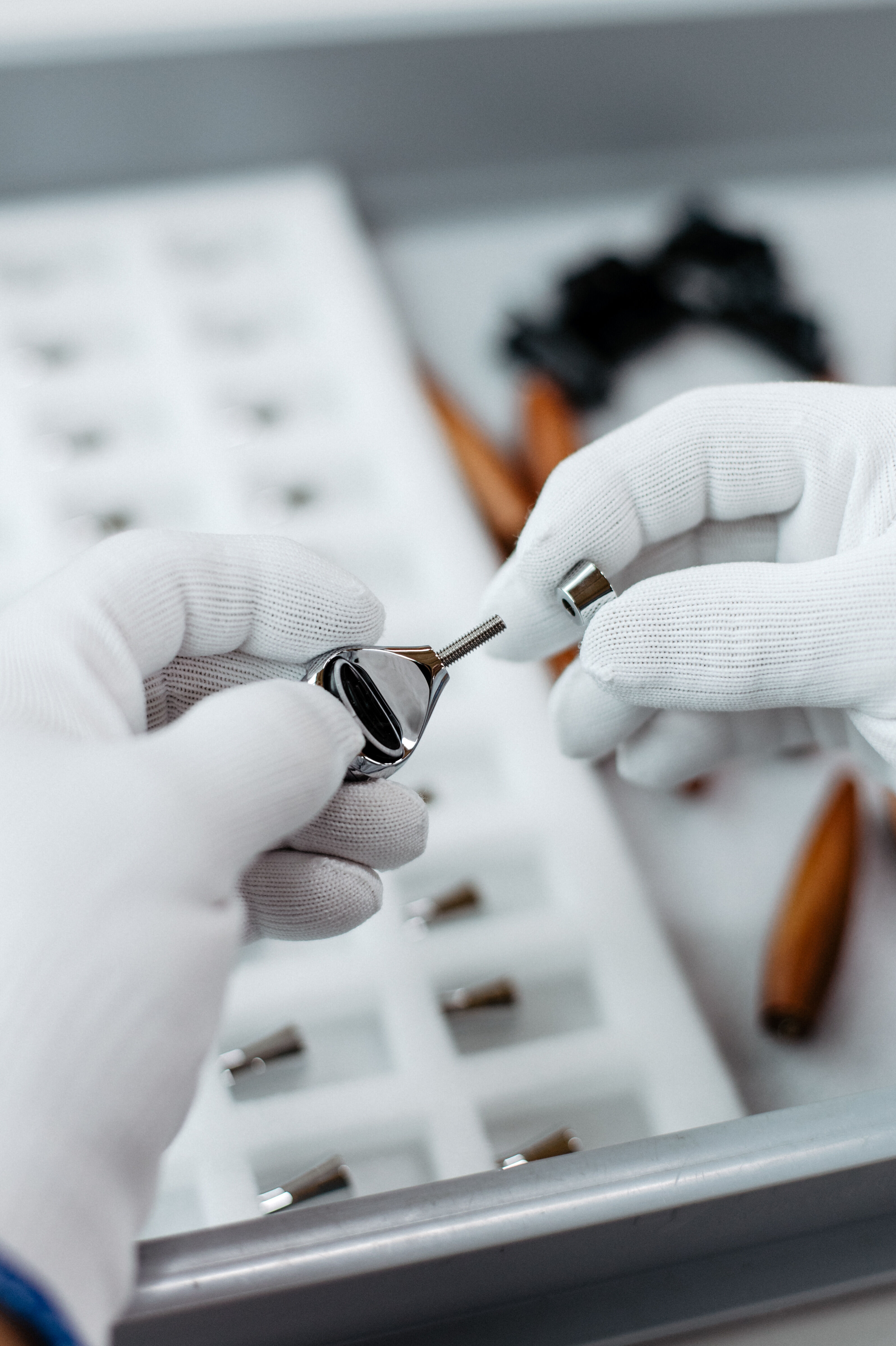 Zwei Hände mit weißen Handschuhen halten ein zerlegtes, glänzendes Metallobjekt vor einem Tablett mit weiteren Metallteilen