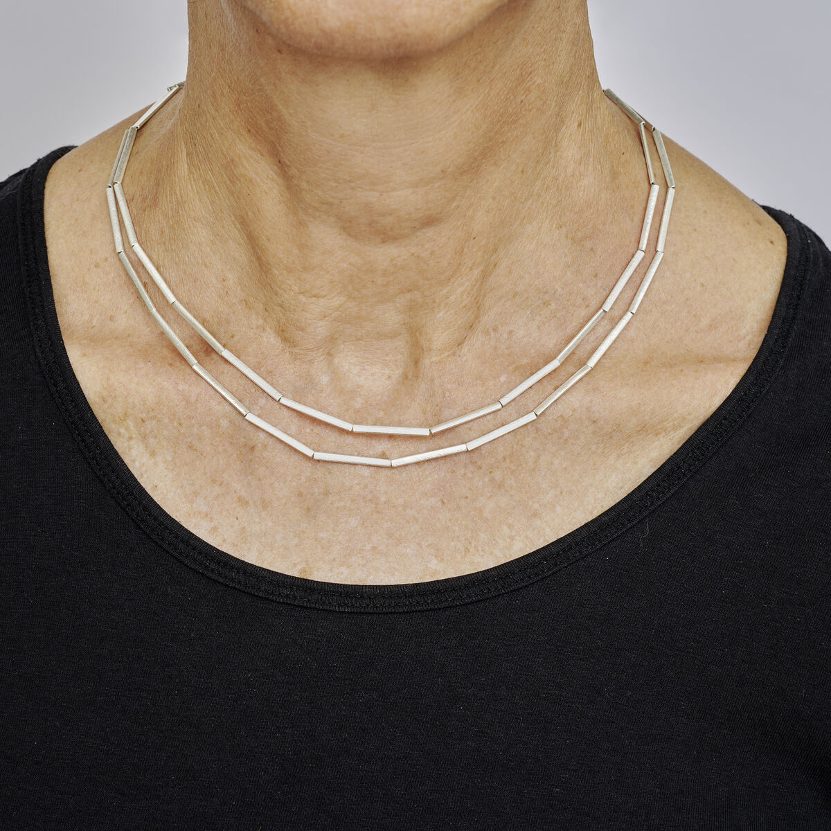 Zweireihige Collier-Halmkette aus Silber an einem Hals einer Frau, präsentiert über einem schwarzen Oberteil, vor neutralem Hintergrund. Die Kette besteht aus schlichten, stäbchenförmigen Silbergliedern und wirkt elegant sowie zeitlos.