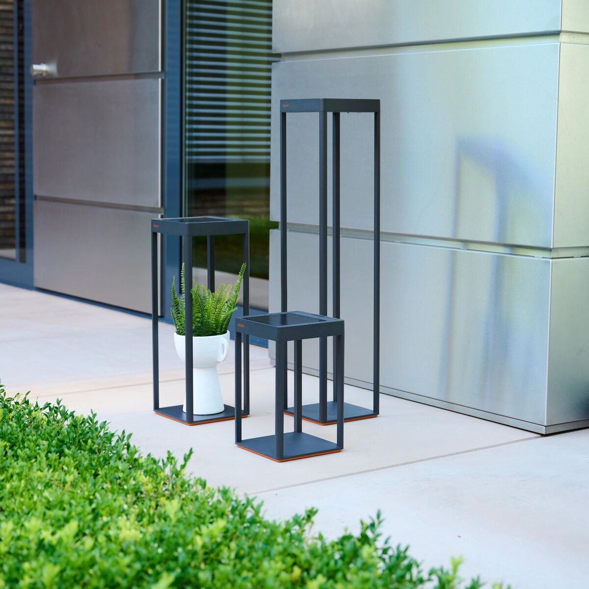 Drei Solarlaternen »WEGA« von Sienagarden in moderner, eckiger Form und anthrazitfarbenem Aluminium stehen dekorativ neben einer weißen Pflanzschale mit Farn vor einem modernen Gebäude. Die Laternen erzeugen stilvolle Akzente auf Terrasse oder Balkon.