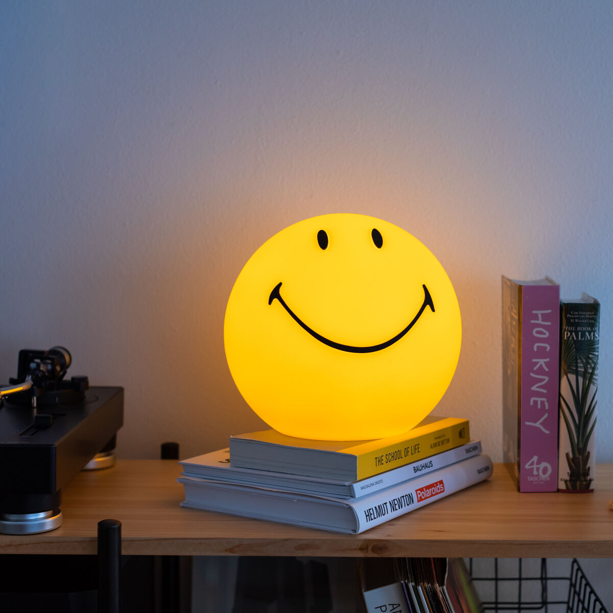 Eine runde, gelbe LED-Lampe in Form eines Smiley-Gesichts steht beleuchtet auf einem Holzregal. Daneben liegen Bücher und eine dekorative helle Skulptur, im Hintergrund eine Schallplatte und weiße Wand. Die Szene strahlt eine freundliche und moderne Wohnatmosphäre aus.