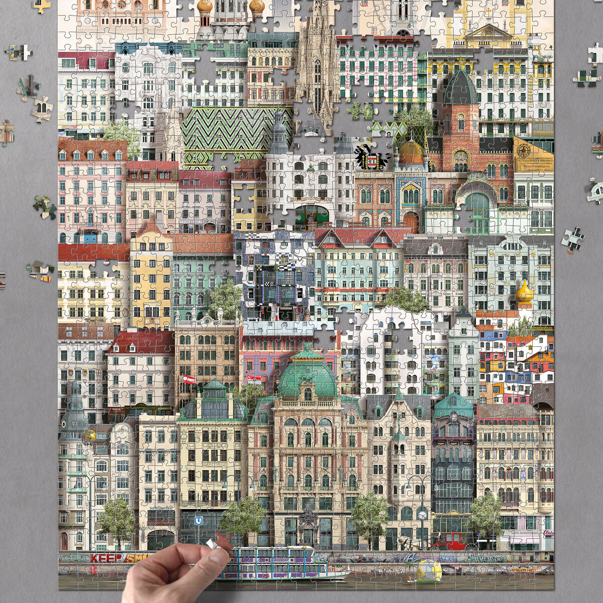 Farbintensives 1000-Teile-Puzzle mit einer detailreichen Illustration ikonischer Gebäude der Stadt Wien. Mehrere architektonische Stile und berühmte Wahrzeichen sind schichtartig übereinander angeordnet. Einzelne Puzzleteile liegen noch um das fertige Bild verteilt. Das Puzzle befindet sich auf einem grauen Untergrund, eine Hand platziert ein Teil am unteren Rand. Besonders geeignet als Geschenk für Städtefans und Familien.