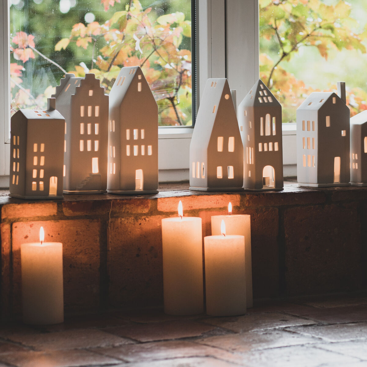 Eine Reihe von weißen Lichthäusern mit Giebeldach und verschiedenen Hausformen stehen auf einer Fensterbank. Mehrere Kerzen in Weißtönen leuchten und ergänzen die gemütliche Szene. Im Hintergrund sind grüne Blätter zu sehen.