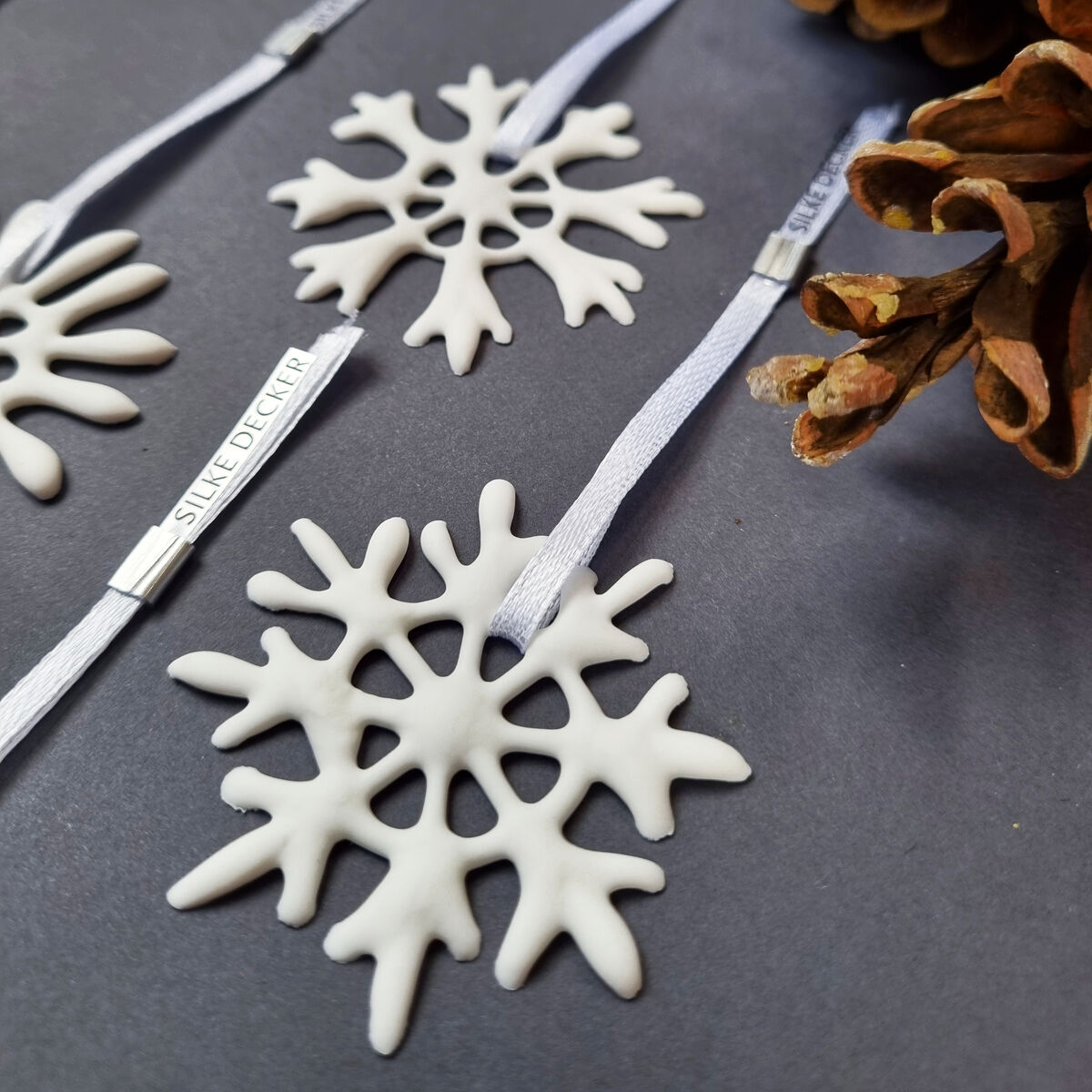 Mehrere weiße Schneeflockenanhänger aus Keramik liegen auf einer grauen Fläche und sind mit weißen Bändern versehen. Einige größere Kiefernzapfen sind zwischen den Anhängern arrangiert. Die Anhänger tragen ein Etikett mit dem Schriftzug SILKE DECKER.