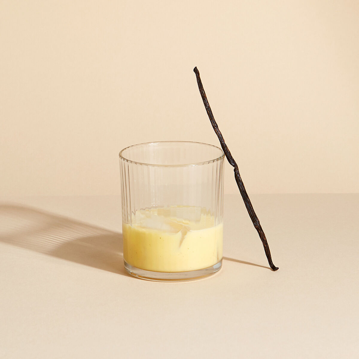 Ein Glas mit cremigem, gelbem Eierlikör steht auf hellem Untergrund. Am Glas lehnt eine lange, braune Vanilleschote. Die Szene ist minimalistisch und hell ausgeleuchtet, Umgebung und Hintergrund sind beigefarben gehalten.