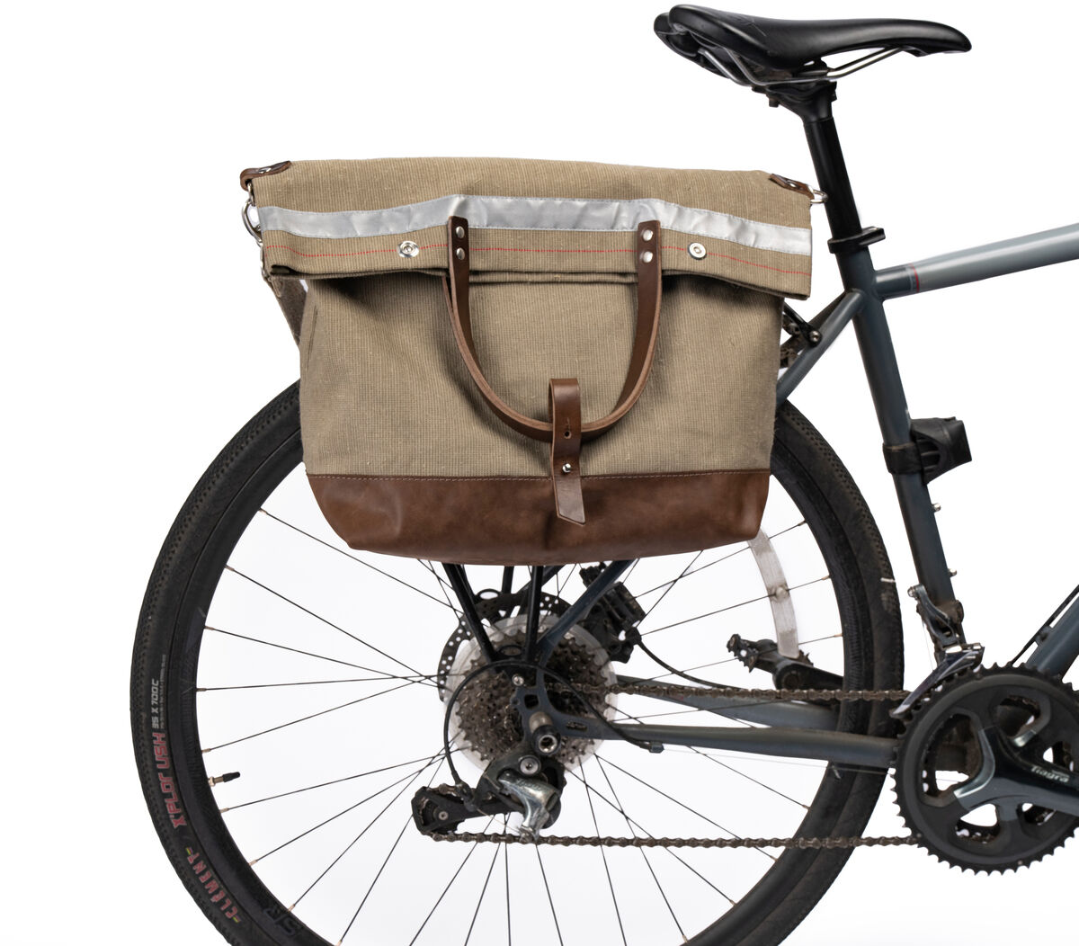 Eine beige und braune Fahrradtasche aus robustem Material ist am Gepäckträger eines Fahrrads befestigt. Das Fahrrad hat einen schwarzen Sattel und einen schwarzen Rahmen. Das Rad des Fahrrads ist ebenfalls sichtbar.