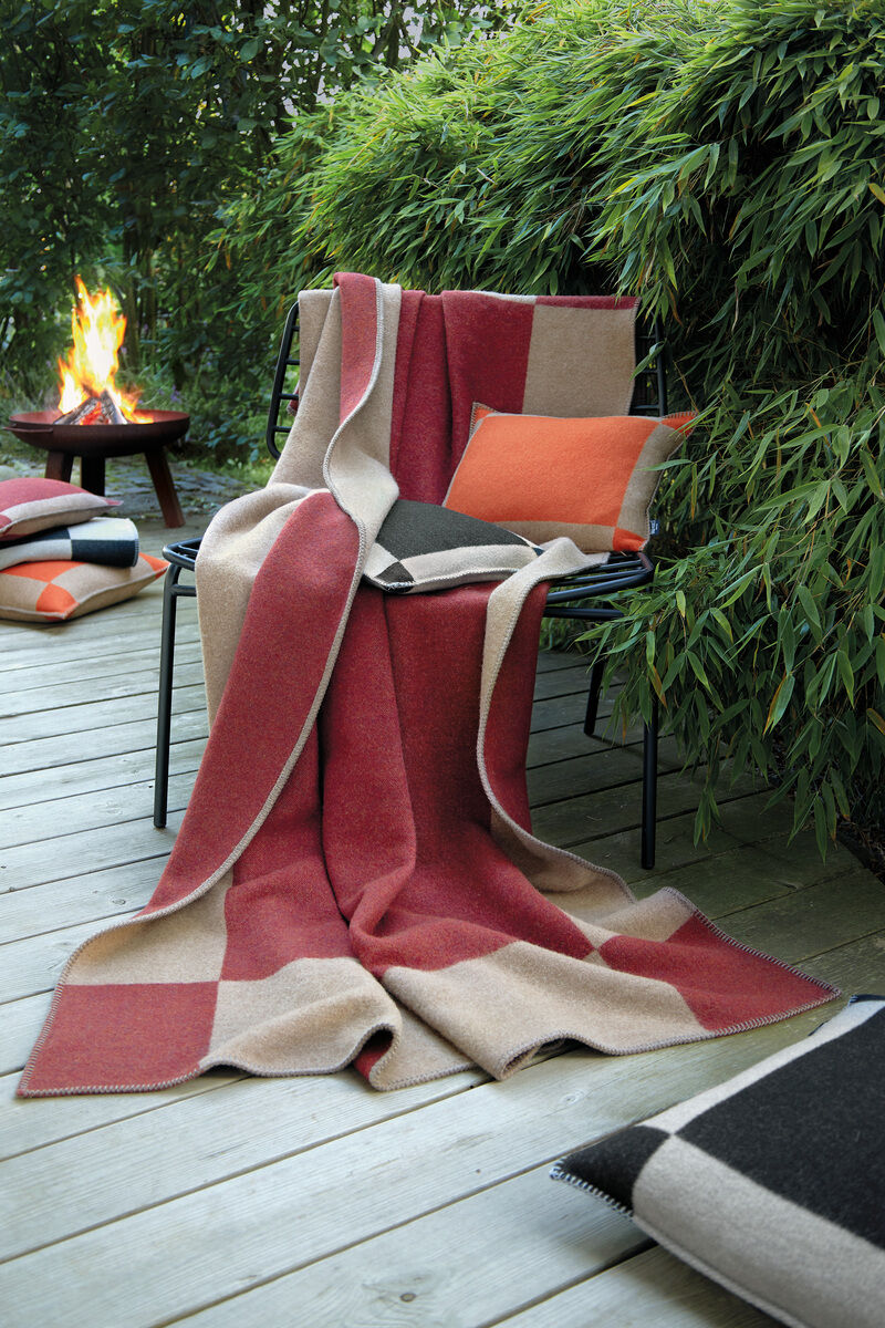Gemütliche Decke »Manchester« in Rot, Beige und Schwarz auf einem Stuhl. Im Hintergrund ein Feuer und Bambussträucher. Ein orangefarbenes Kissen auf dem Stuhl und ein weiteres Kissen am Boden ergänzen die Szene. Perfekt für drinnen und draußen, insbesondere in der kühleren Jahreszeit.
