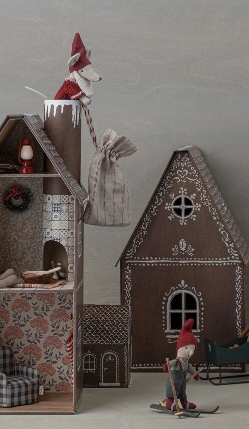 Eine Szene mit zwei niedlichen Weihnachtsmann-Mausfiguren. Eine Maus sitzt auf einem Holzhaus und hält eine gestreifte Geschenktüte, während die andere auf Skiern steht. Die Häuser sind dekoriert, eines davon wie ein Lebkuchenhaus bemalt, und die Figuren sind in Rot und Weiß gekleidet. Die Szene ist festlich dekoriert und erinnert an ein gemütliches Weihnachtsmärchen.