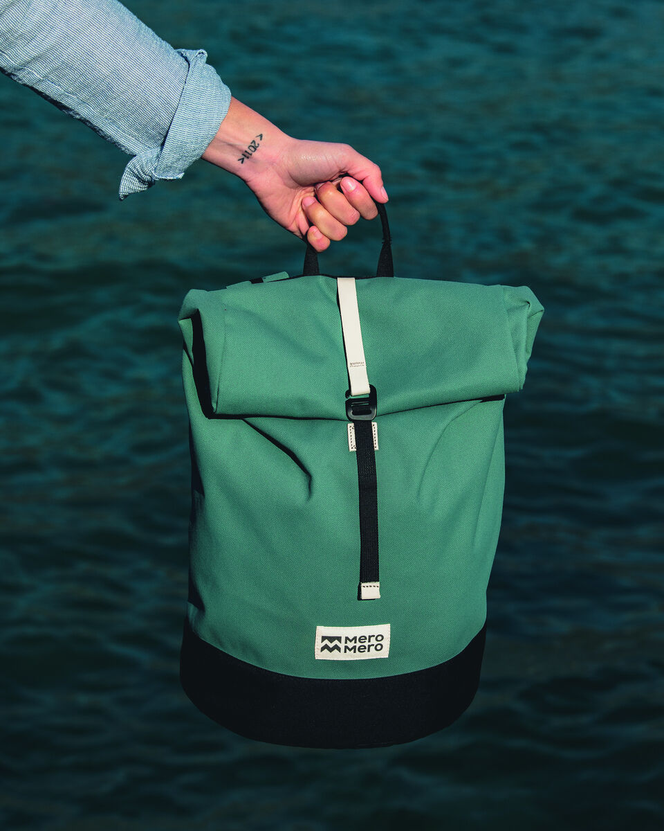 Eine grüne Fahrradtasche namens »Annecy« wird von einer Hand gehalten, die sich vor einem blauen Hintergrund aus Wasser befindet. Die Tasche hat schwarze und weiße Akzente und trägt das Logo 'Mero Mero'.