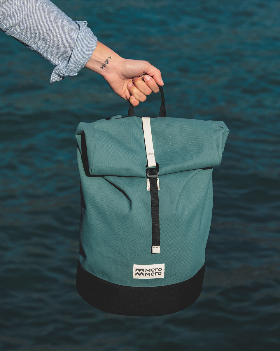 Eine grüne Fahrradtasche namens »Annecy« wird von einer Hand gehalten, die sich vor einem blauen Hintergrund aus Wasser befindet. Die Tasche hat schwarze und weiße Akzente und trägt das Logo 'Mero Mero'.