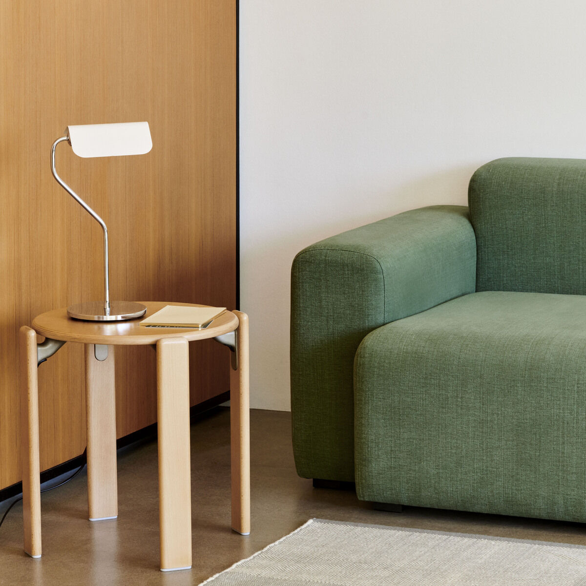 Eine moderne Tischlampe steht auf einem kleinen Holztisch neben einem grünen Sofa in einem minimalistischen Wohnzimmer. An der Wand hängt eine Uhr. Der Raum hat helle Wände und einen Holzakzent.
