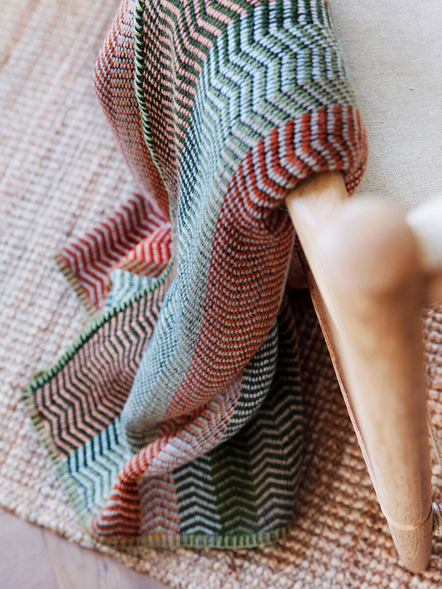 Lammwolldecke »Fri«, geripptes und gemustertes Design in Beige, Orange, Grün und Braun, über eine Holzlehne drapiert, auf einem geflochtenen Teppich liegend.