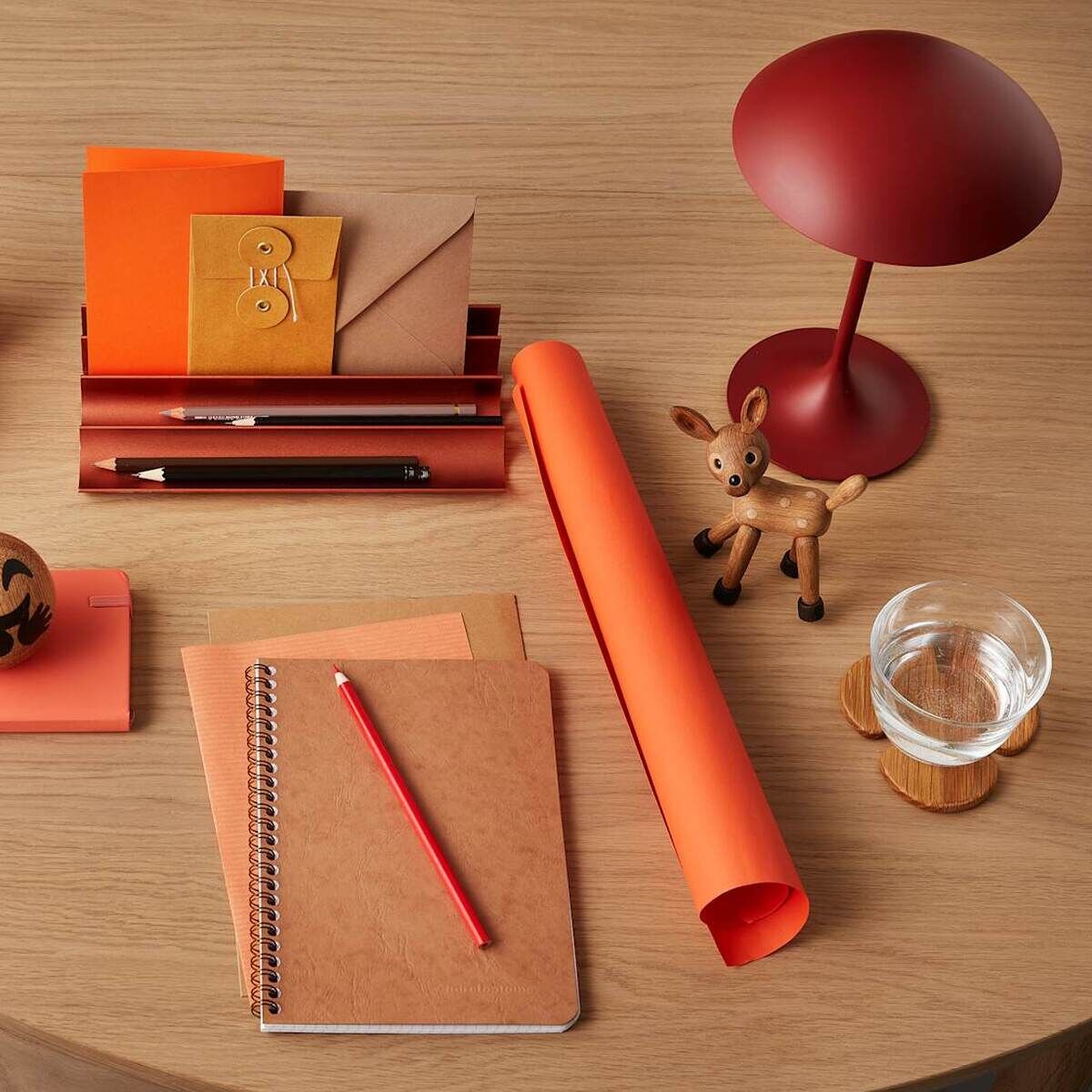 Auf einem runden Holztisch stehen verschiedene Büromaterialien in Rot-, Braun- und Orangetönen, darunter eine kleine Holzfigur in Form eines Rehs, ein Glas Wasser, eine rote Lampe und Notizbücher. Die Szene wirkt ordentlich, natürlich und kreativ gestaltet.