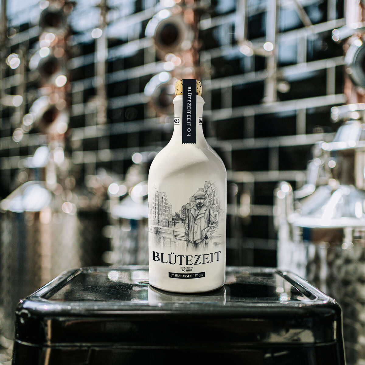 Eine weiße Designerflasche des ZEIT-Jubiläumsedition Gins »BLÜTEZEIT« steht auf einem schwarzen Tischchen in einem modernen Destillerieumfeld. Im Hintergrund sind glänzende Destillationsgeräte mit schwarzen Fliesen zu sehen.
