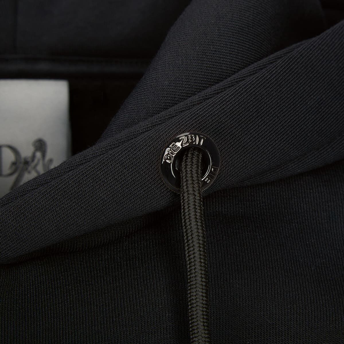 Nahaufnahme einer schwarzen ZEIT-Kollektion »ZEIT Backprint« Hoodie-Kapuze mit einem Metallösen-Detail und Logo.