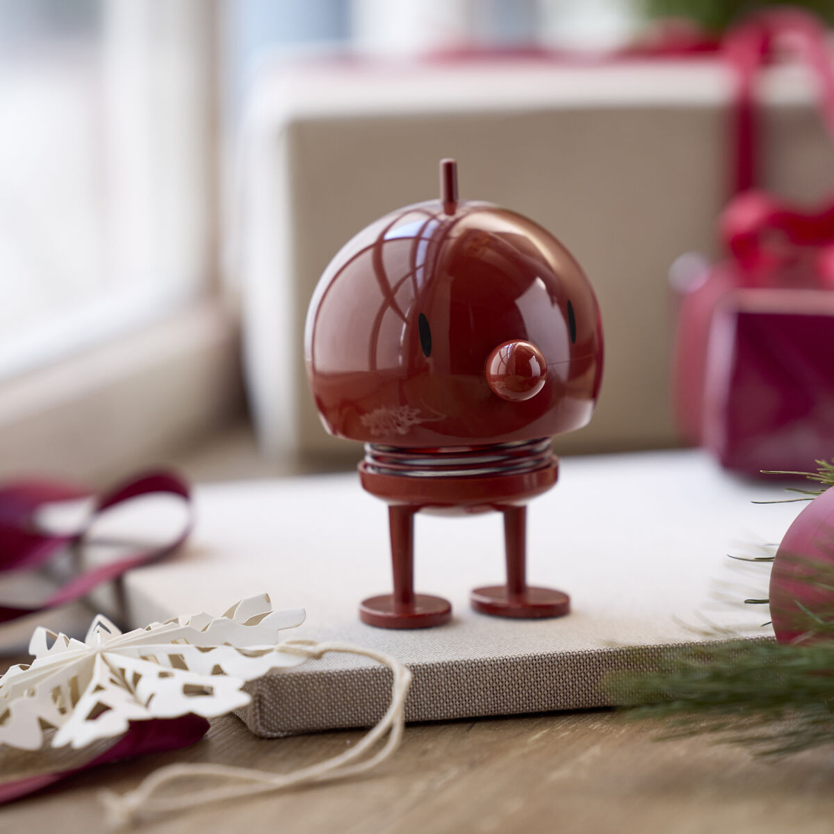 Der Hoptimist Bumble Maroon von F&H GmbH ist eine bordeauxrote, stilisierte Kunststofffigur mit runder Form und Spiralfeder auf zwei Beinen. Die Szenerie zeigt Geschenkboxen, Tannenzweige sowie weiße Holz-Schneeflockendeko auf einem Holztisch in festlicher Atmosphäre.