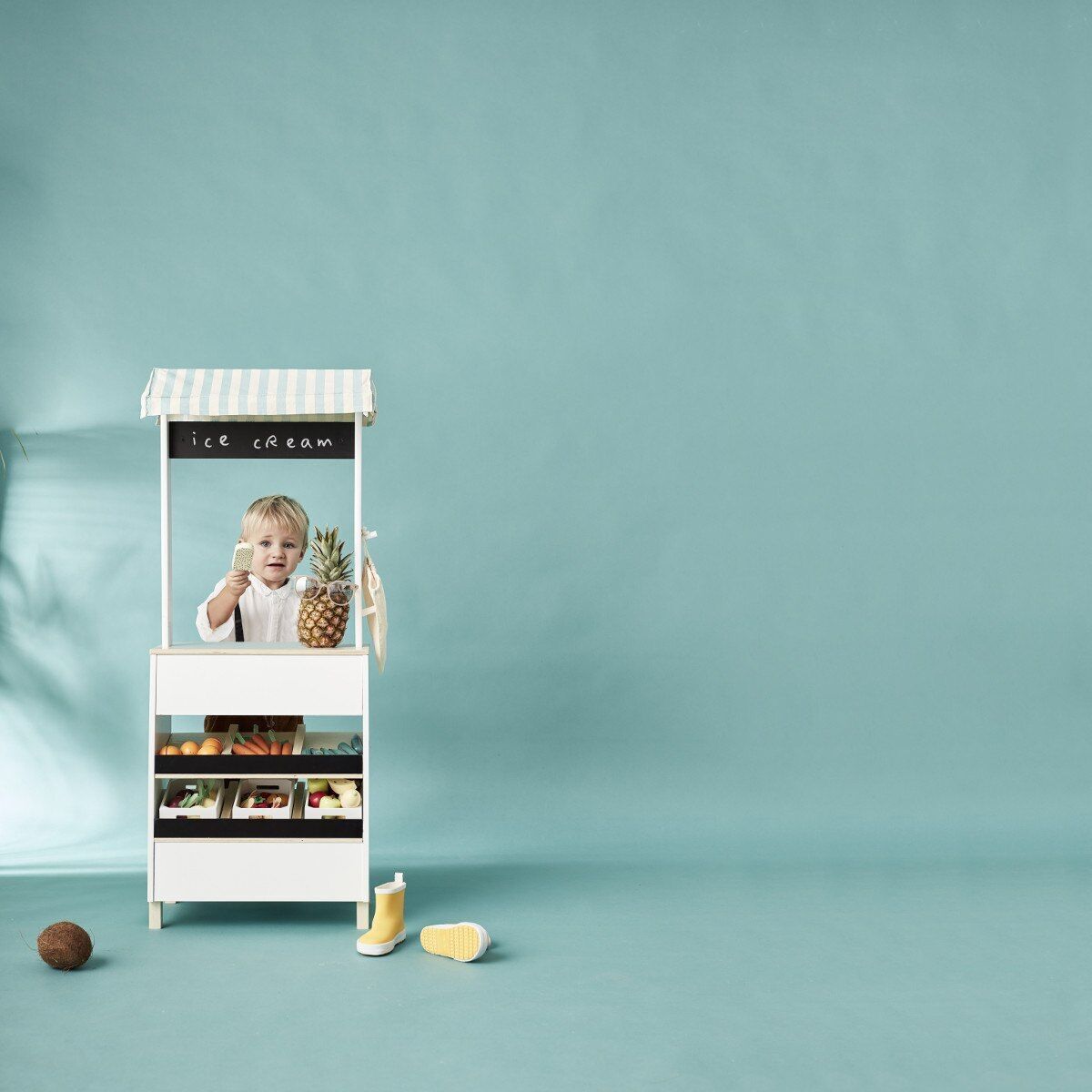 Ein weißer Marktstand für Kinder mit Sonnendach auf hellblauem Hintergrund. Vor dem Stand sitzt ein Kind und hält eine Ananas. Im Marktstand sind verschiedene Spielzeugfrüchte und -gemüse ausgestellt. Links neben dem Stand liegt ein Ball, rechts eine halbe Orange.