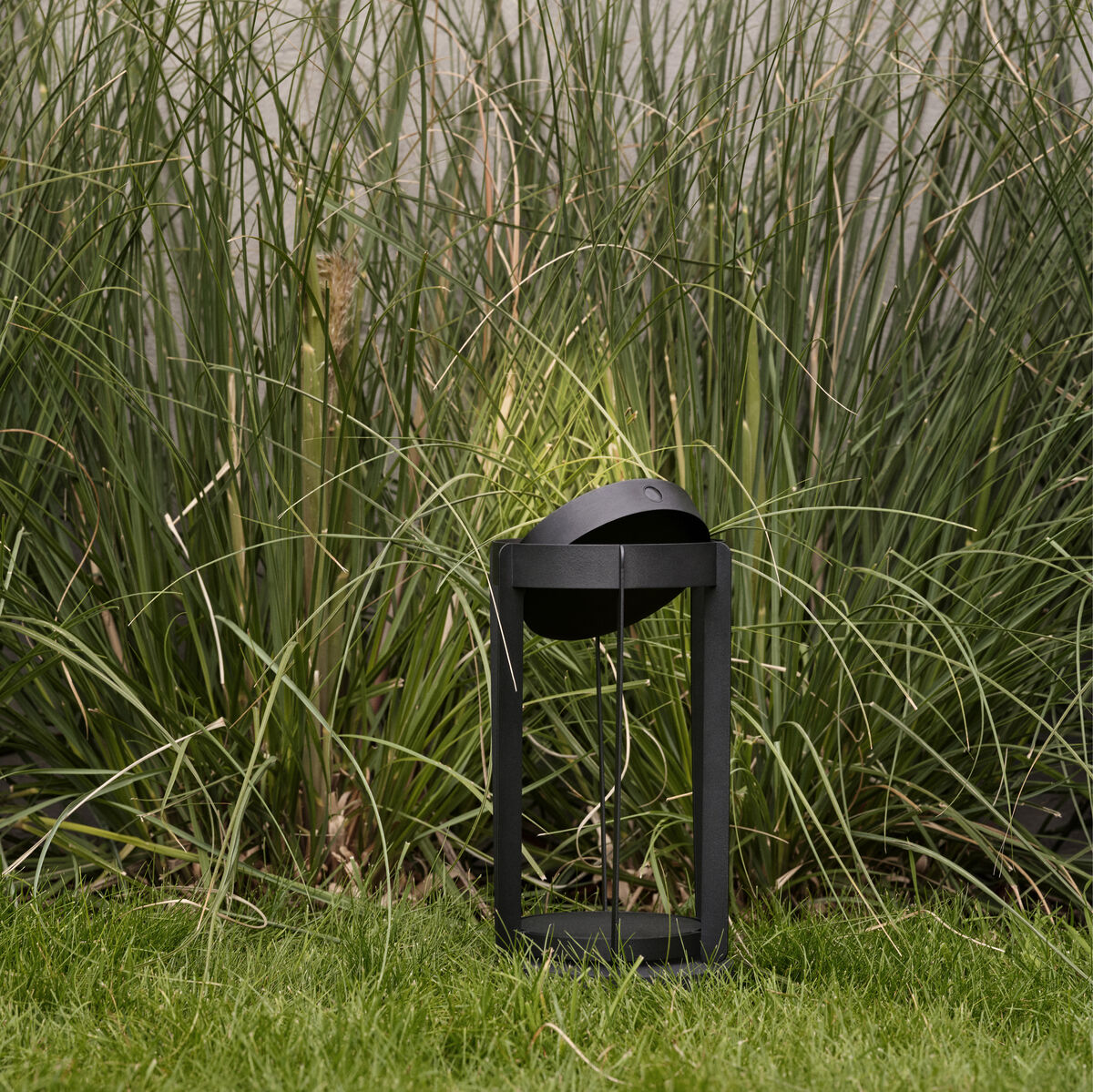 Eine schwarze LED-Laterne »Firefly« steht auf einem grünen Rasen, im Hintergrund sind hohe Gräser zu sehen.