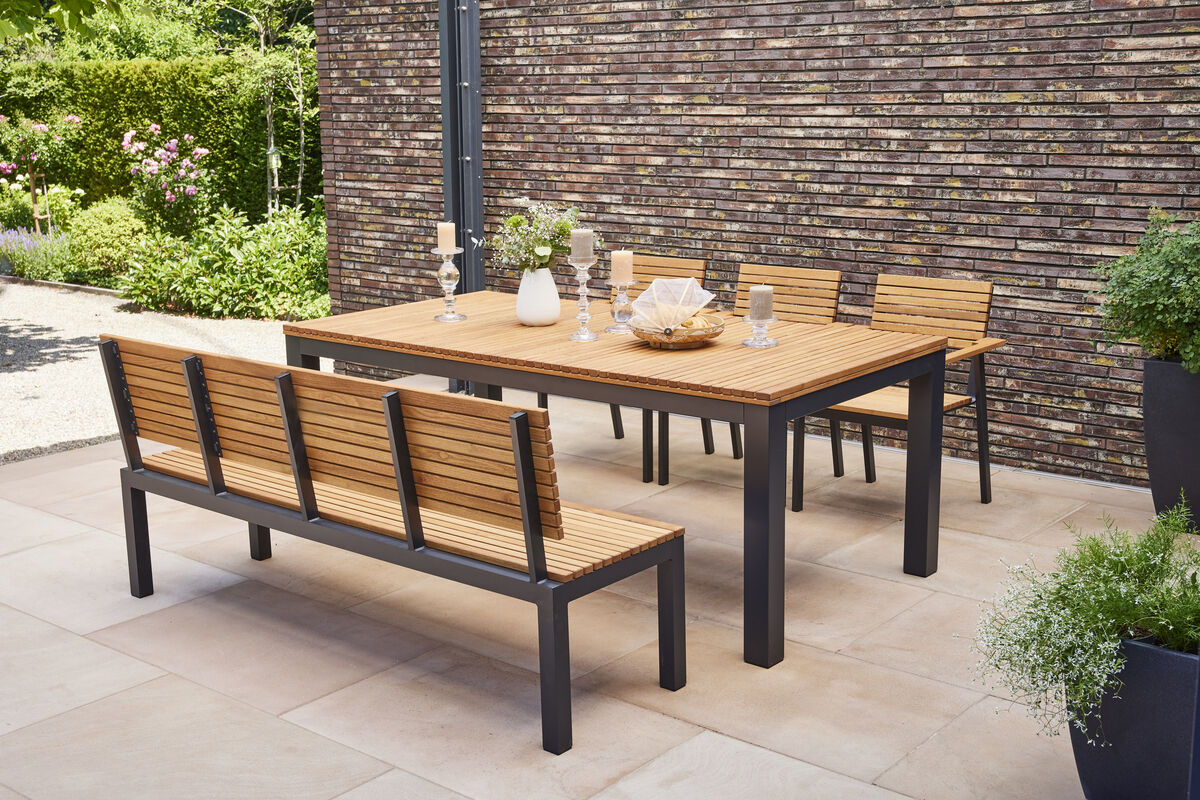 Ein moderner Esstisch aus Holz und Metall mit passender Gartenbank und Stühlen, platziert auf einer Terrasse mit natürlicher Gartenumgebung im Hintergrund.