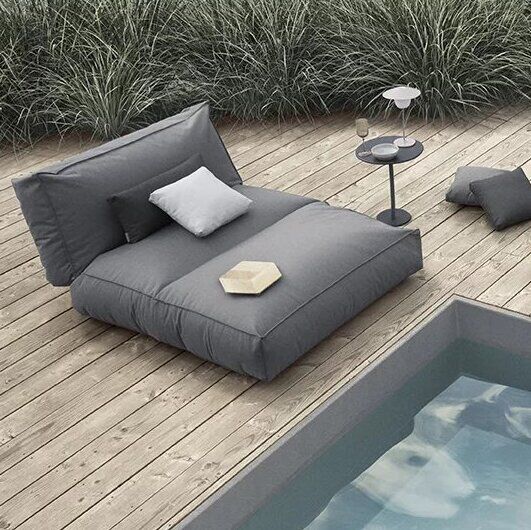 Ein Outdoor-Bett »STAY« 120 x 190 cm, umgeben von anderen luxuriösen Gartenmöbeln auf einer Holzterrasse neben einem Pool in Wassernähe. Die Möbel sind in Grautönen gehalten und verleihen dem Bereich ein modernes und entspanntes Ambiente.