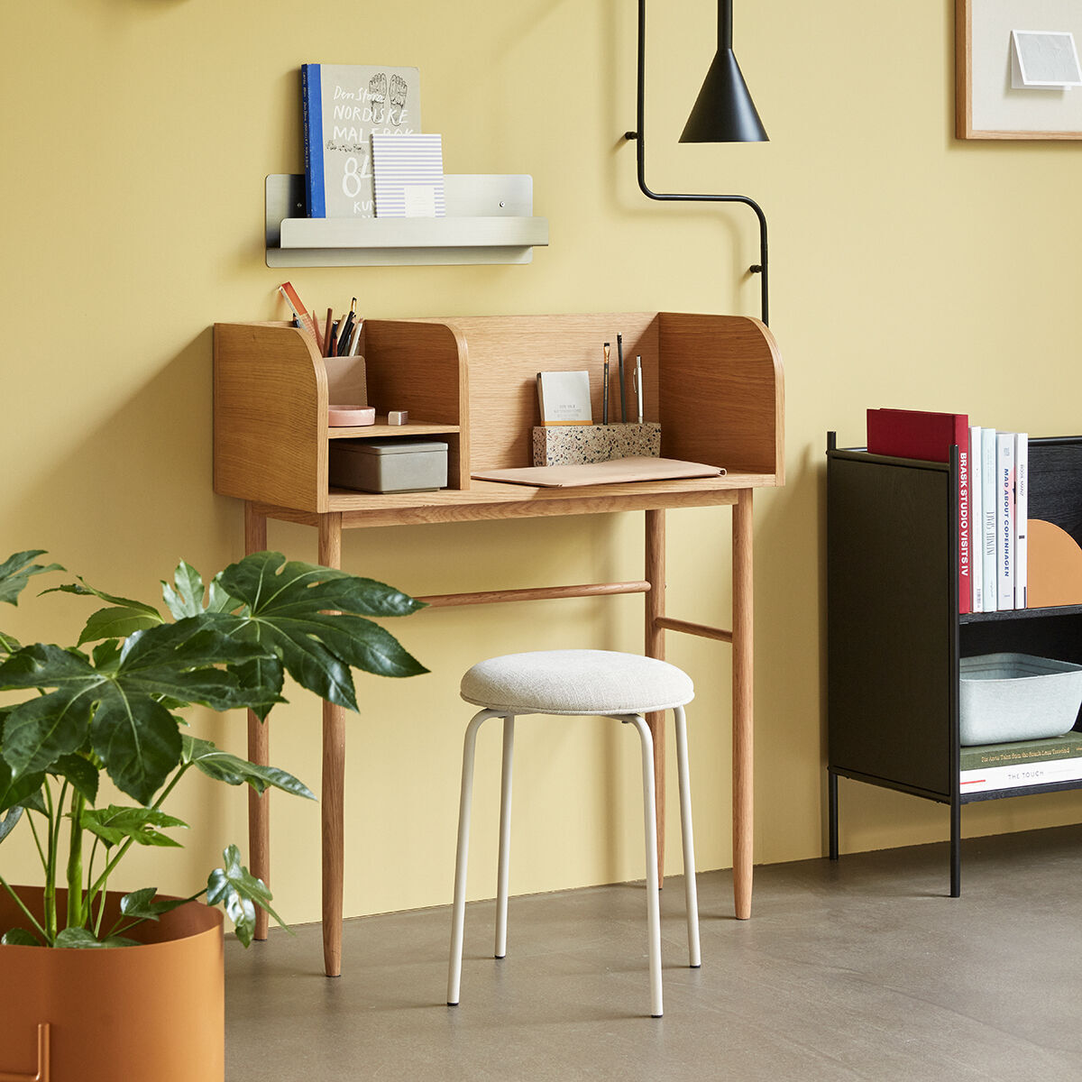 Ein moderner Schreibtisch aus Eichenholz befindet sich in einem hellen Raum mit einem Hocker. Der Schreibtisch ist minimalistisch und funktional gestaltet, mit Ablagen und einem Regal darüber. Ein Pflanzenkübel steht links vom Schreibtisch, während rechts Bücherregale und Dekorationsgegenstände stehen. Die Wand ist gelb gestrichen, und es gibt Wandlampen für zusätzliche Beleuchtung.