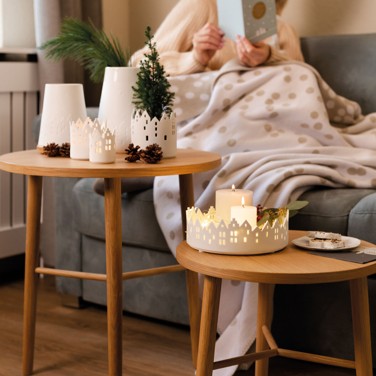 Ein gemütliches Wohnzimmer mit einer Frau, die in eine Decke gehüllt auf einem Sofa sitzt und eine Karte liest. Auf zwei nebeneinander stehenden Holztischen stehen weiße Stadtschalen, Vasen mit Tannenzweigen, Kerzen und weihnachtliche Dekoration im skandinavischen Stil. Die Szene vermittelt eine warme, festliche Atmosphäre.