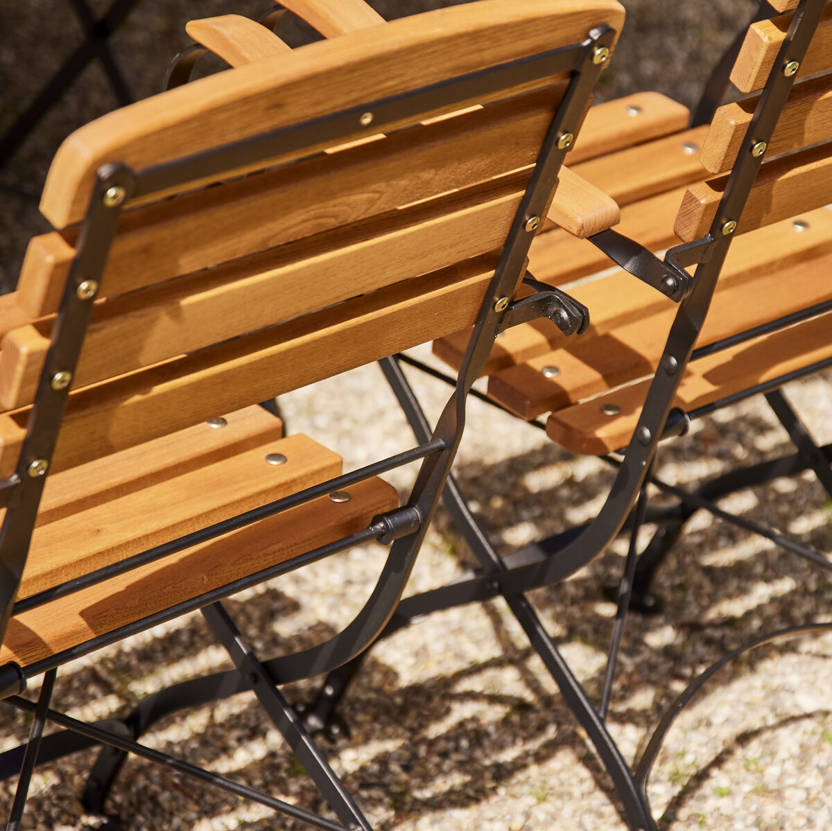 Zwei klappbare Gartenstühle aus Holz mit Stahlrahmen stehen auf einem Kiesboden in der Sonne.