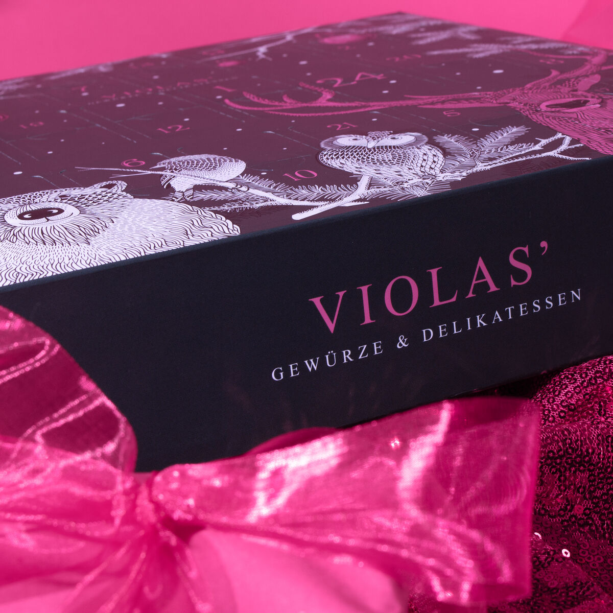 Ein luxuriöser Adventskalender von Violas in Rosa, Schwarz und Weiß, verziert mit einem großen rosa Band, bietet eine Auswahl an Gewürzen und Delikatessen als Geschenk zur Adventszeit.