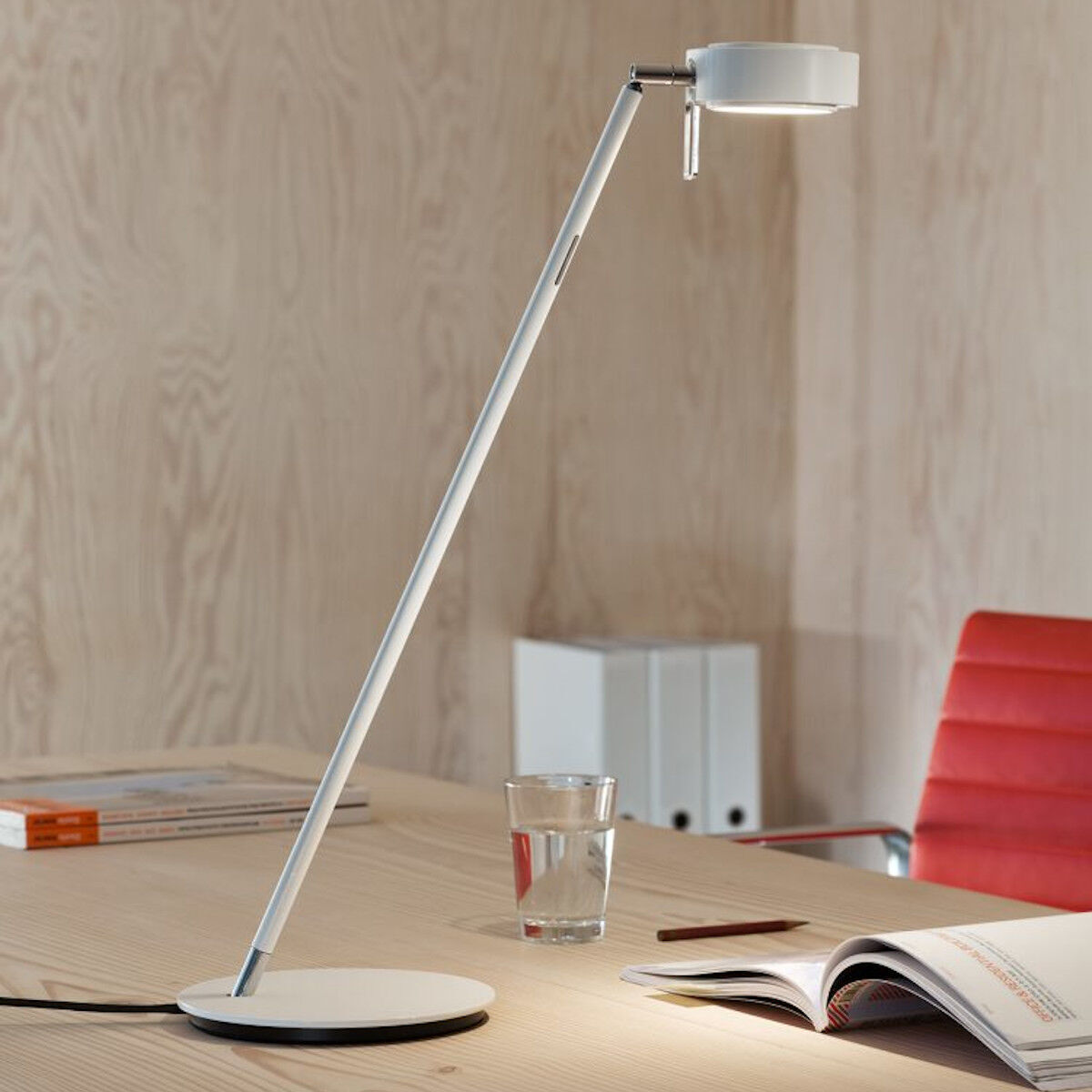 Eine moderne Tischleuchte namens »Pure« ist auf einem Schreibtisch platziert. Die Leuchte hat ein minimalistisches Design mit einem schlanken, weißen Korpus und einer runden Basis. Auf dem Tisch befinden sich ein roter Stuhl, Bücher und eine geöffnete Zeitschrift. Die Wand im Hintergrund ist in einem hellbeigen Farbton gehalten.