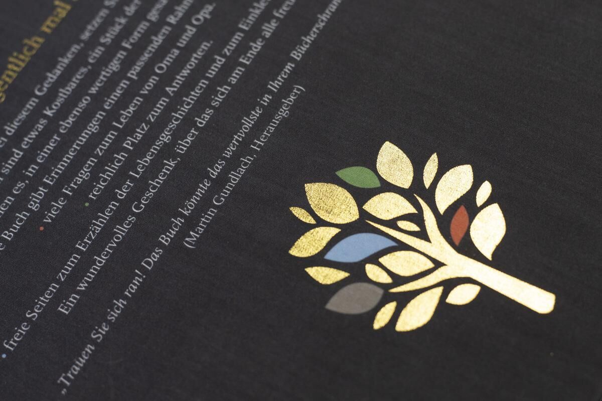 Ein schwarzes Erinnerungsbuch mit dem Titel 'Erzähl mir dein Leben - Leinenausgabe', verziert mit einer goldenen Prägung eines stilisierten Baumes. Die Blätter des Baumes sind in verschiedenen Farben wie Grün, Blau und Rot gestaltet. Der Einband des Buches ist aus Leinen.