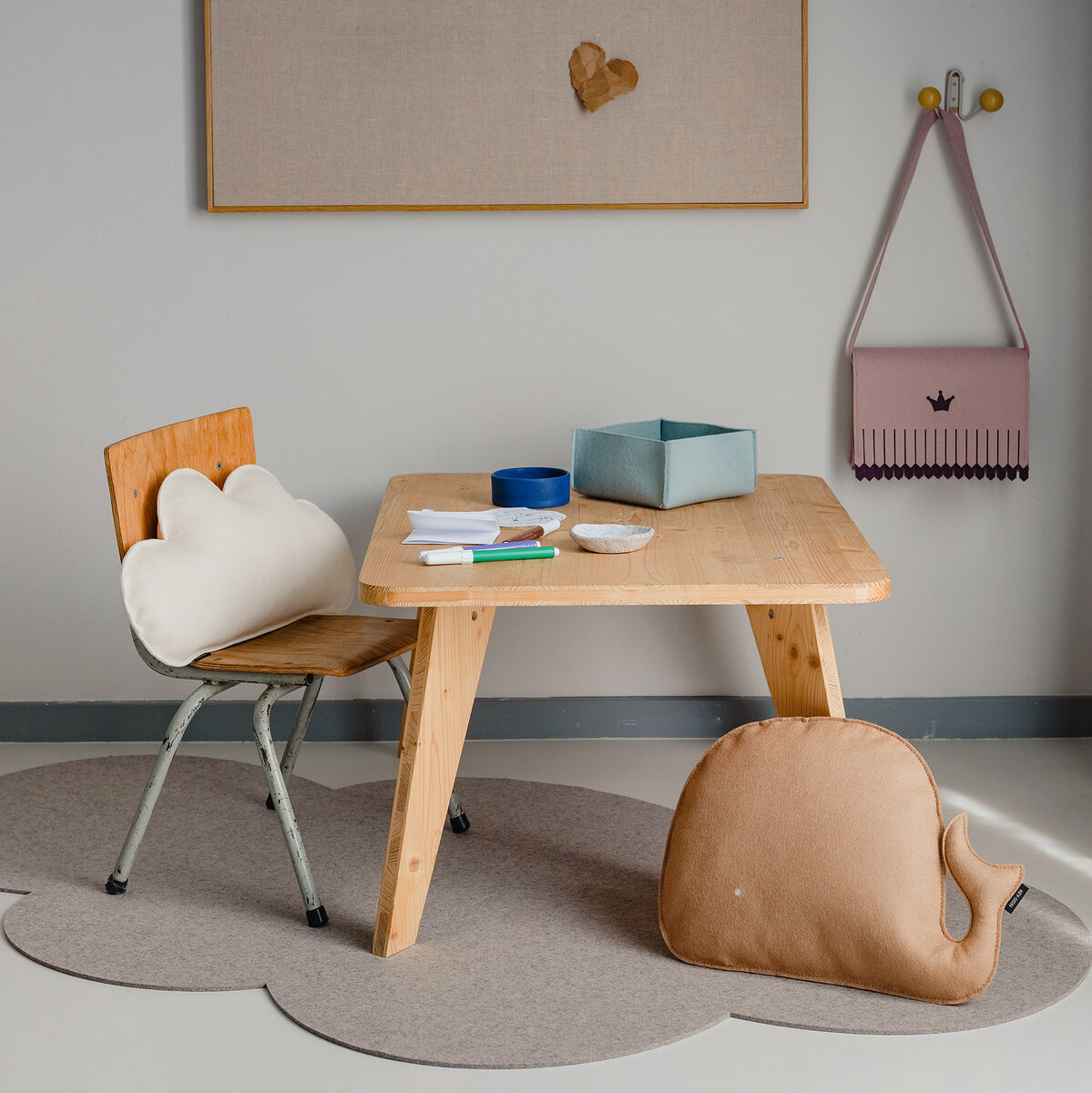 Gemütliches Kinderzimmer mit einem Wolkenkissen auf einem Holzstuhl vor einem kleinen, rechteckigen Holztisch. Verschiedene Accessoires und Dekorationen sind im Raum verteilt, darunter ein geformter Spielteppich, ein Aufbewahrungskorb und weitere dekorative Elemente.