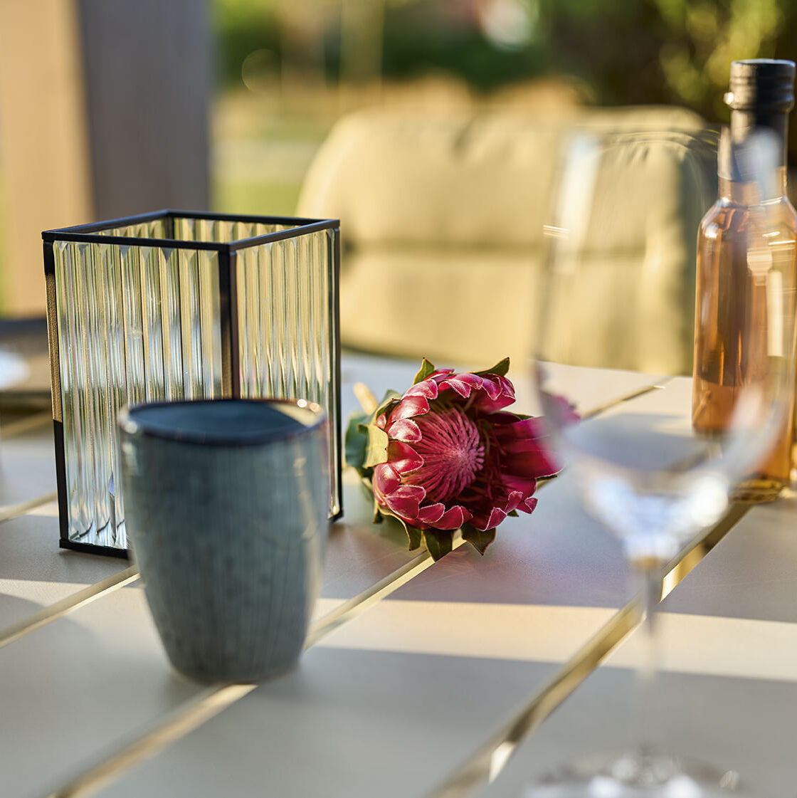 Ein stilvoller Esstisch (Tisch »VAPIO« 180x90 cm, basic) auf einer Terrasse, dekoriert mit Kerzen, Weinflaschen, einer Blume und Gläsern. Im Hintergrund unscharf erkennbar sind Gartenstühle und Pflanzen.