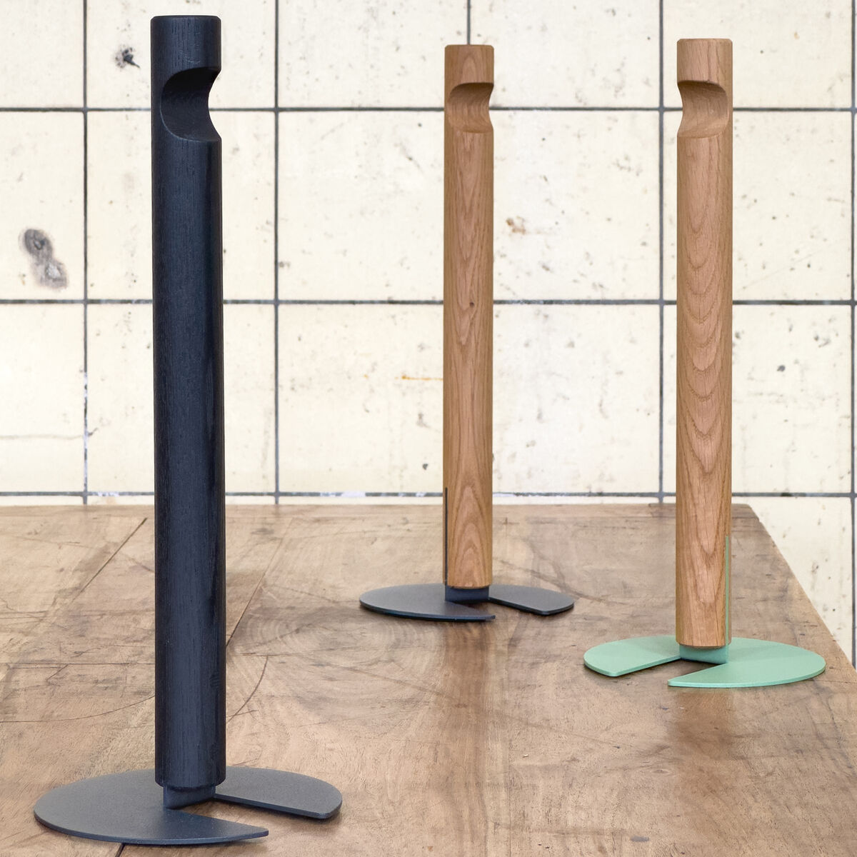 Drei Küchenrollenhalter GIVE der Marke finbeaux von Christoph Schoen stehen auf einem Holztisch vor einer gefliesten Wand. Zwei Modelle sind aus Naturholz mit verschiedenfarbigen Bodenplatten (grau, grün), einer ist dunkel mit grauer Bodenplatte, minimalistisch und modern gestaltet.