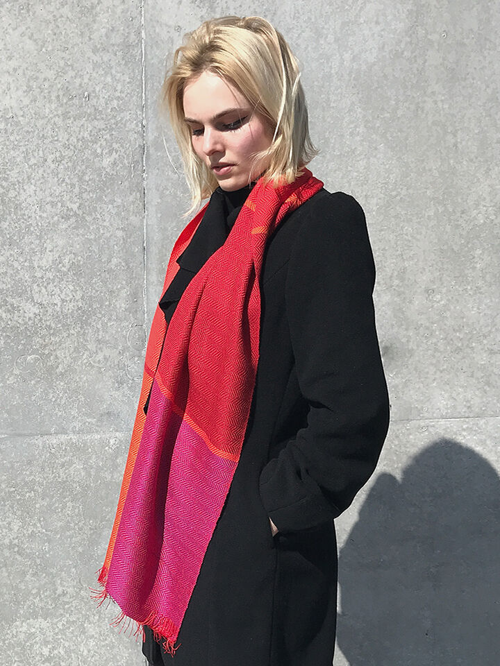 Eine Frau trägt einen Seidenschal in Rot, Orange und Pink vor einer grauen Wand. Sie trägt einen schwarzen Mantel.