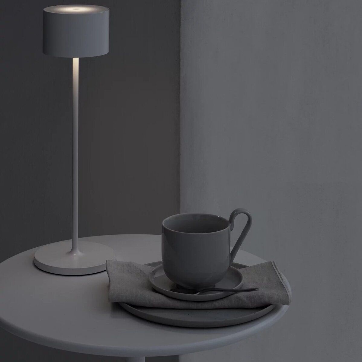Eine minimalistisch gestaltete, graue LED-Tischleuchte steht auf einem runden Tisch zusammen mit einem grauen Becher und einer grauen Serviette. Die Umgebung ist in gedeckten Grautönen gehalten.