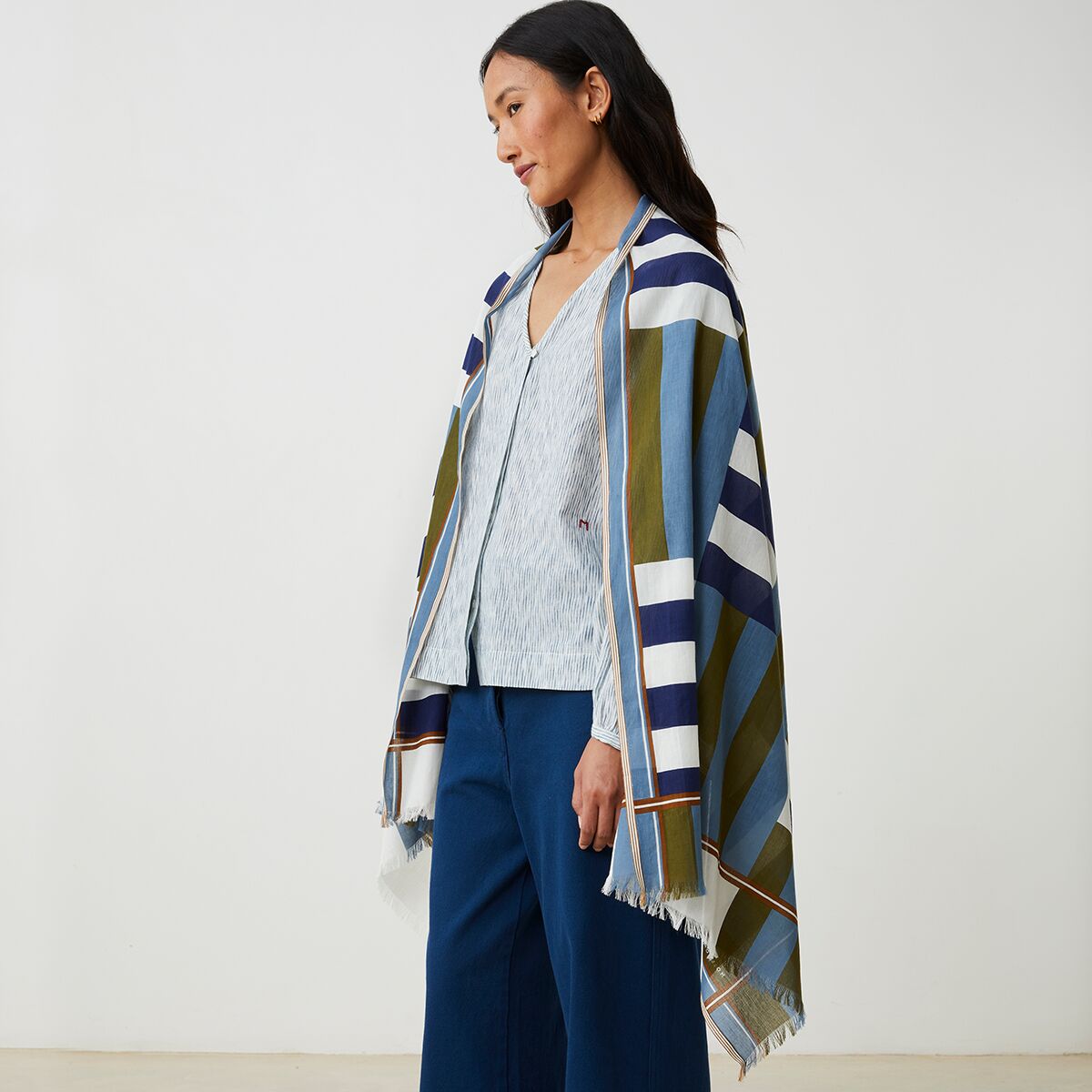 Eine Frau trägt den Baumwollschal No. 771 von MOISMONT über Schultern und Oberkörper. Der Schal ist breit, besteht aus 100% Baumwolle und zeigt breite Streifen in Blau, Weiß, Dunkelblau und Olivgrün. Er hat kurze Fransen an den Enden. Die Kleidung darunter ist schlicht in Blau- und Beigetönen.