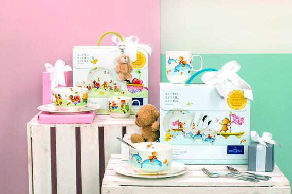 Das Bild zeigt das Kindergeschirr-Set »Hungry as a Bear« mit Tassen, Schüsseln und Tellern mit bunten Tiermotiven. Zwei Geschenkverpackungen sind vorhanden, verziert mit weißen Schleifen und einem kleinen Stoffbären. Der Hintergrund ist pastellrosa und mintgrün, und ein weißer Holzstuhl dient als Unterlage. Die Atmosphäre ist fröhlich und kindgerecht.