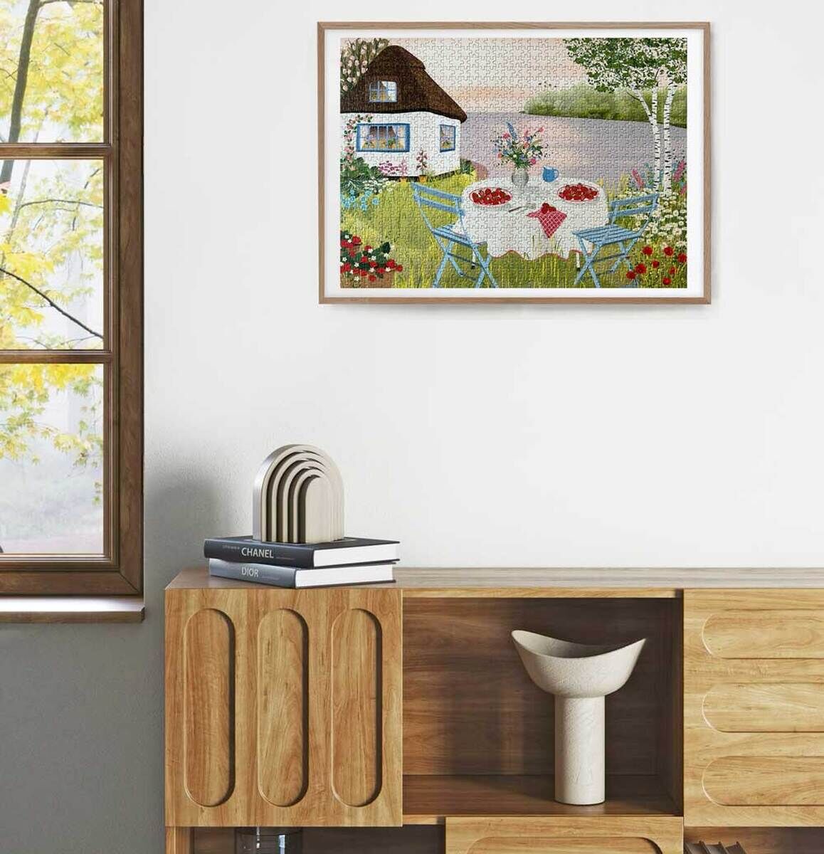 Ein gerahmtes Puzzle mit dem Titel 'Süße Erdbeeren', das an einer weißen Wand hängt. Es zeigt eine idyllische Gartenszene mit einem kleinen Haus, grünen Pflanzen und blauen Gartenmöbeln. Neben dem Puzzle ist ein Fenster mit gelblichen Herbstblättern sichtbar. Darunter befindet sich ein schlichter Holztisch mit Büchern und dekorativen Elementen.