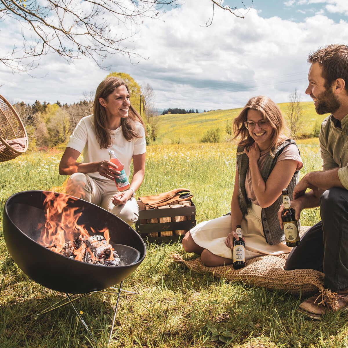 Eine Gruppe von drei Personen sitzt im Freien auf einer Wiese und genießt ein gemütliches Beisammensein neben einer brennenden Feuerschale. Im Hintergrund ist eine natürliche Landschaft mit Bäumen und einer Hängesessel-Schaukel zu sehen. Der Himmel ist teils bewölkt und die Atmosphäre wirkt entspannt und leicht fröhlich.