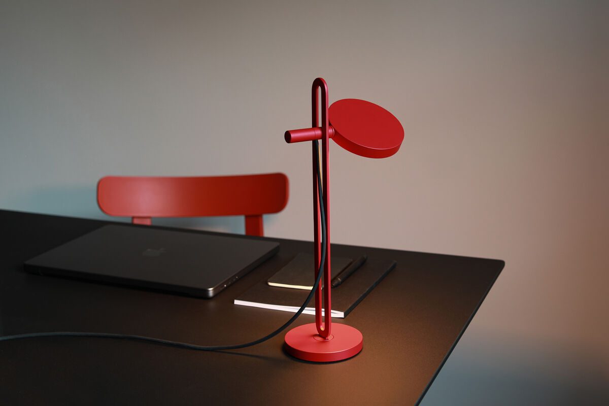Eine moderne rote Tischlampe mit einem minimalistischen Design steht auf einem schwarzen Schreibtisch. Die Lampe hat ein schlankes Metallgestell und ist an einen schwarzen Stromkabel angeschlossen. Im Hintergrund sieht man einen roten Stuhl und einen geschlossenen schwarzen Laptop.